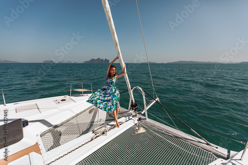 Slender tall charming model posing while standing on toes on a large white yacht, holding a handrail with her hands on the background of the sea.