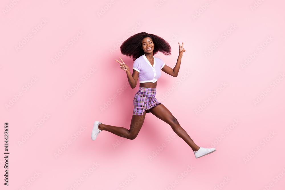 Full length body size view of beautiful trendy cheerful girl running jumping showing v-sign isolated over pink pastel color background