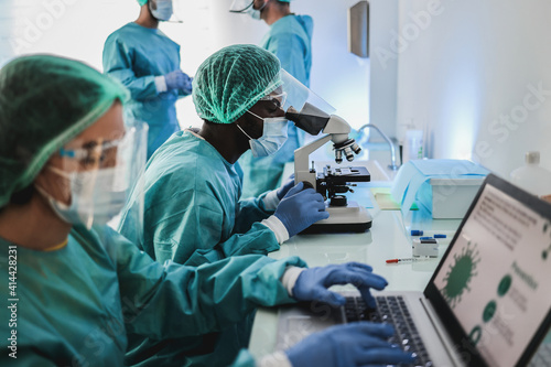 Multiracial medical scientist in hazmat suit working with microscope and laptop computer inside modern laboratory hospital - Focus african man face