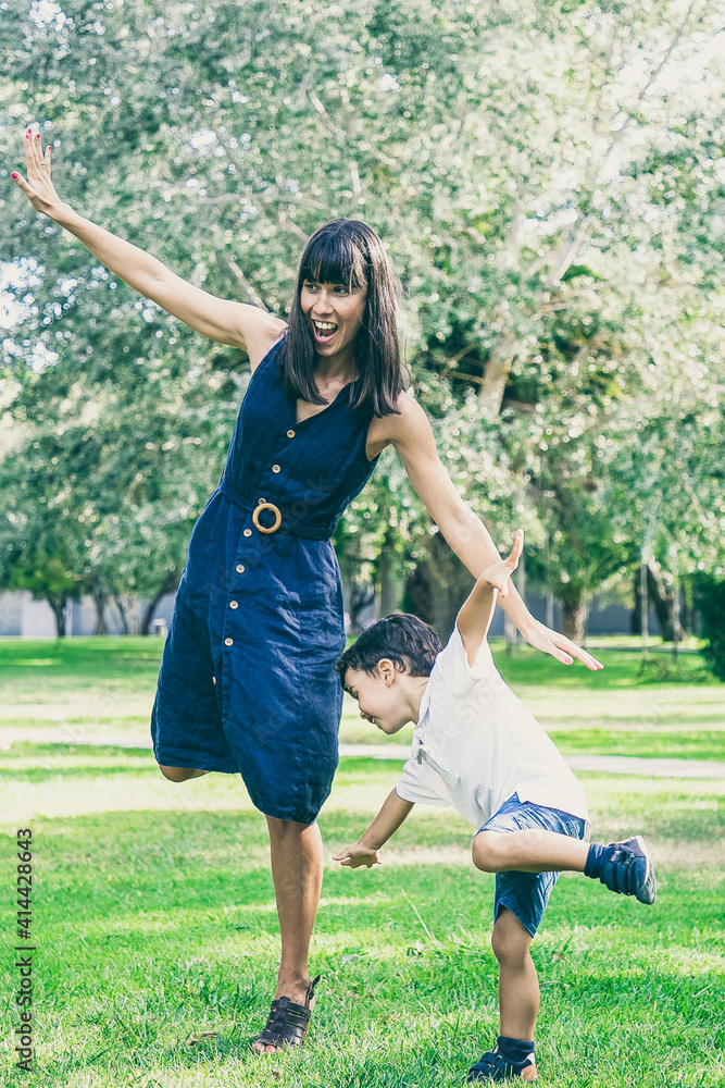 Excited mom and little son playing active games outdoors, standing and balancing on one leg, doing funny exercises in park. Family outdoor activity and leisure concept Excited mom and little son playing active games outdoors, standing and balancing on one leg, doing funny exercises in park. Family outdoor activity and leisure concept