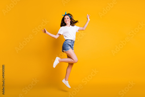 Full length body size photo of playful girl jumping looking at empty space smiling isolated on vibrant yellow color background