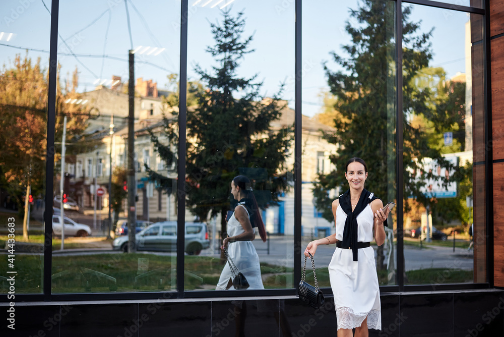 Young brunette girl with red pony tail, wearing stylish white silk dress,running jumping in front of glass building, holding phone, smiling.Business woman on lunch break.Romantic female urban portrait