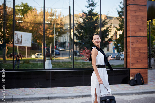 Young brunette girl with red pony tail, wearing stylish white silk dress, walking in front of glass building, holding phone. Pretty business woman on lunch break. Romantic female urban portrait.