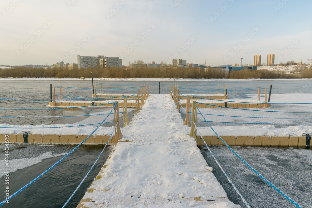 Modular boat pier on the river. Winter season, snow. Background - city, buildings, sky, trees.