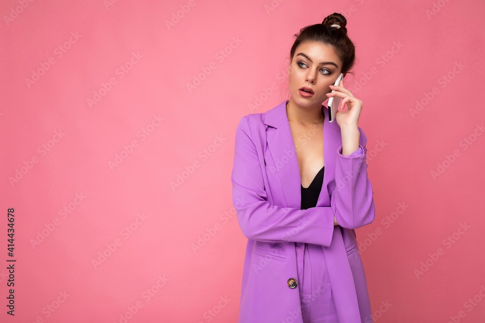 Photo of dissatisfied beautiful young woman wearing purple suit talking on the mobile phone isolated over pink background looking to the side Photo of dissatisfied beautiful young woman wearing purple suit talking on the mobile phone isolated over pink background looking to the side