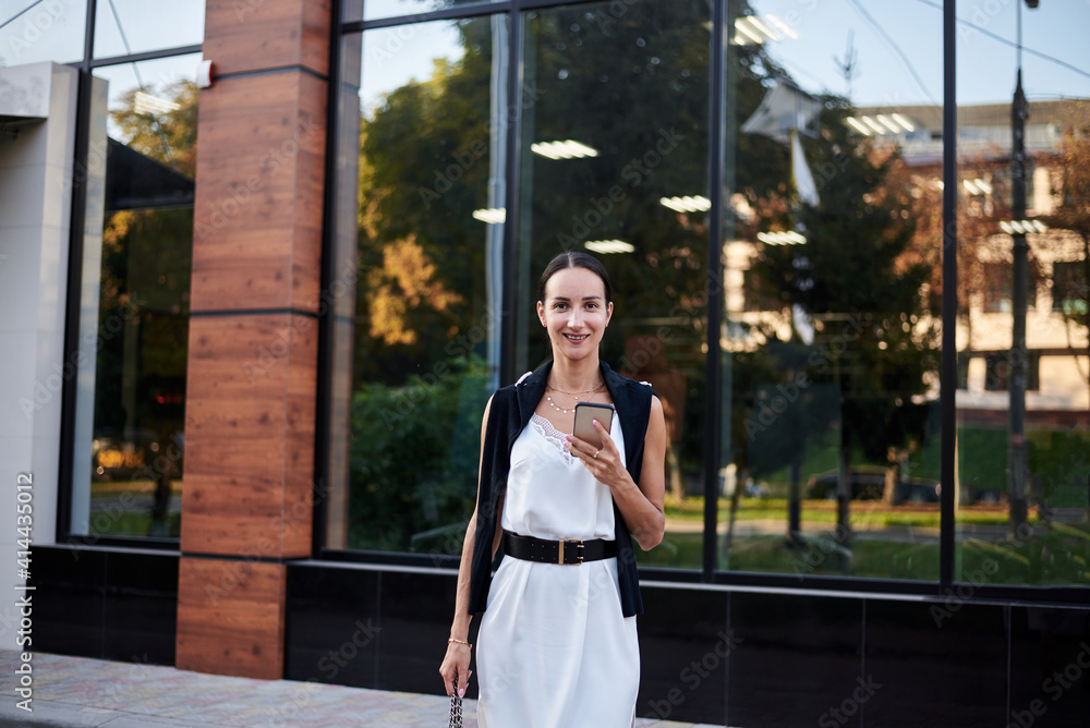 Young brunette girl with red pony tail, wearing stylish white silk dress, walking in front of glass building, holding phone. Pretty business woman on lunch break. Romantic female urban portrait.