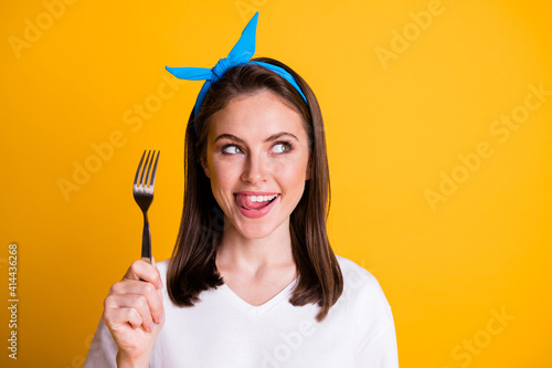 Photo of charming lady hold fork lick teeth look empty space wear blue headband white t-shirt isolated yellow color background