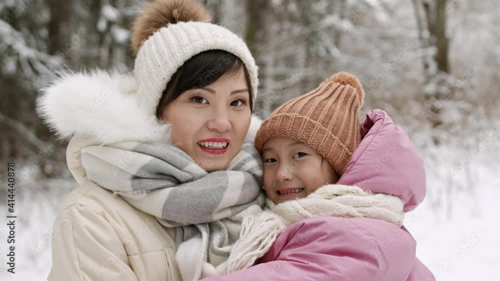 Medium close-up POV of cheerful Asian mother and child wearing puffer jackets, hats with pompoms and scarves posing in snowy winter park. Daughter kissing mom, then looking on camera Medium close-up POV of cheerful Asian mother and child wearing puffer jackets, hats with pompoms and scarves posing in snowy winter park. Daughter kissing mom, then looking on camera