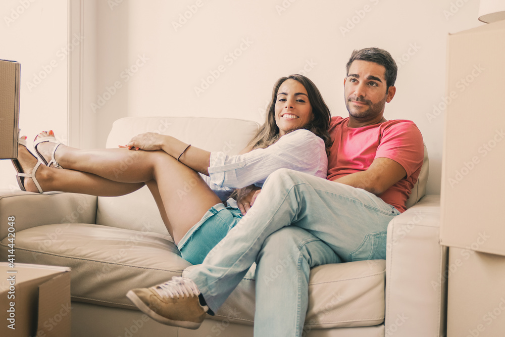 Cheerful Latin young couple sitting on couch among cardboard packages in new apartment, looking at camera and smiling. Moving or new home concept Cheerful Latin young couple sitting on couch among cardboard packages in new apartment, looking at camera and smiling. Moving or new home concept
