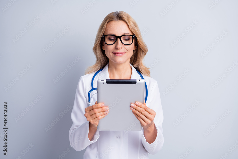 Photo of charming shiny mature lady doctor wear white coat spectacles looking modern device isolated grey color background Photo of charming shiny mature lady doctor wear white coat spectacles looking modern device isolated grey color background