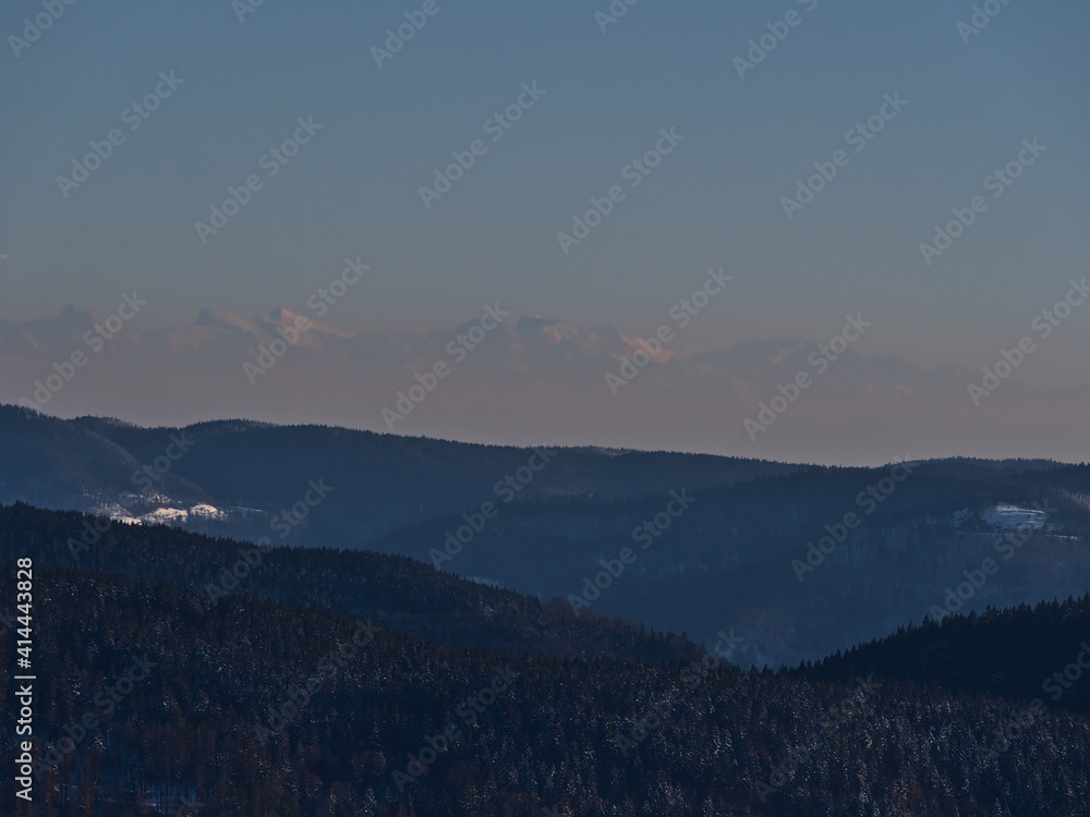 Stunning aerial view of the southern Black Forest hills in winter season with snow-covered landscape viewed from Schauinsland peak, Germany with the silhouettes of the majestic Alps in background.