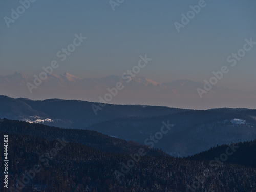 Stunning aerial view of the southern Black Forest hills in winter season with snow-covered landscape viewed from Schauinsland peak, Germany with the silhouettes of the majestic Alps in background.