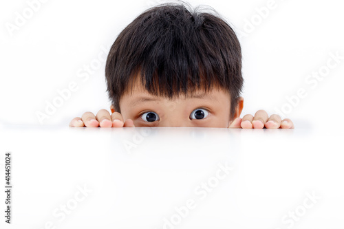 Vászonkép Portrait of an Asian boy peeking through the surface of table isolated on white background