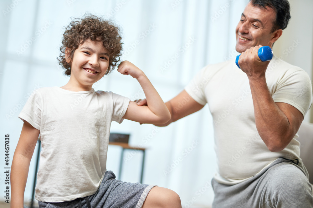 Fototapeta premium Fit boys. Little hispanic boy child smiling at camera showing his biceps while having workout together with his father, exercising at home