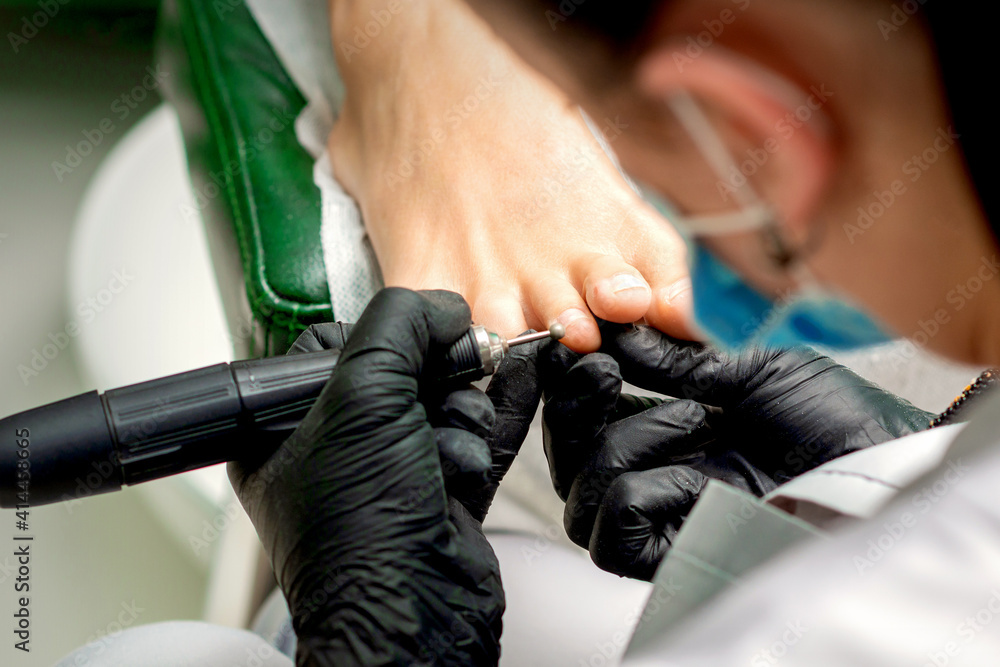 Pedicure master removes corns and cuticles with electric file machine on female toes in a nail