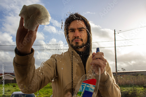 A man with a hoodie is washing a window on the balcony, he sprays detergent on the glass. He holds in one hand a sprays detergent and in another hand he holds a napkin and carefully rubs the glass 
