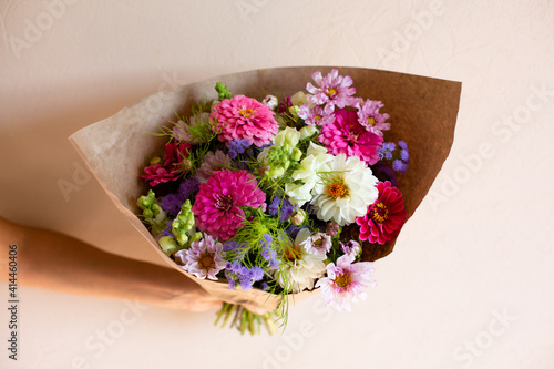 Hand with beautiful bouquet of seasonal flowers on pink background. Pink zinnia, white dahlia, cosmos, purple ageratum, white antirrhineae. Bouquet of seasonal flowers in craft paper. photo