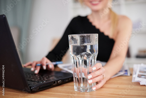 Close-up picture of glass with water on office table in hand of young blond woman, working on laptop, Manager at workplace. Work process in company. Healthy drink consumption concept.Water balance.