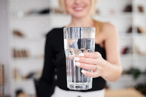 Close-up picture of huge glass of water in hands of young blond woman, standing in office room. Manager at workplace. Work process in company.Healthy drink consumption concept.Water balance.