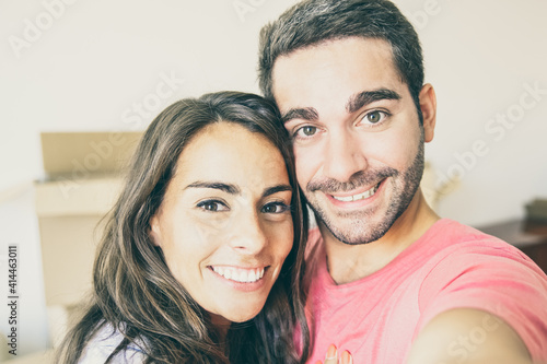 Selfie of happy young couple in their new home, posing with carton boxes in background, holding gadget in hand. Closeup shot. New home and real estate concept