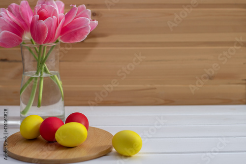 Purple-pink tulips in a transparent vase on a wooden background, next to which are red and yellow Easter eggs, a spring-Easter card, a top view, a place for text