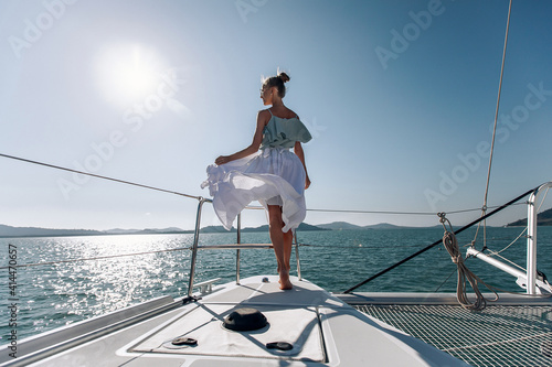 Full-length photo of a skinny young cute model in a long white airy dress, standing on the edge of her yacht against the backdrop of the sea and sun. The sun illuminates the water. Summer concept.
