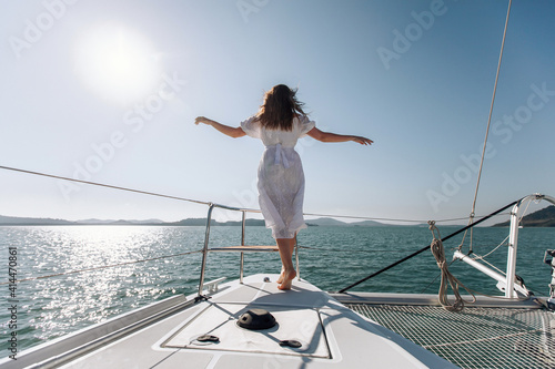 Full-length photo of a young and slender girl in a long white dress posing on the edge of her white yacht, arms outstretched in different directions, enjoying the flow of wind. Concept of freedom. © Semachkovsky 