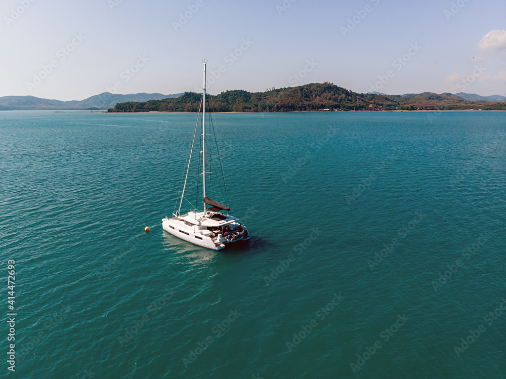 Aerial view of the white luxury yacht floating alone in the exotic turquoise sea near the island filled with forests and at the background of mountains and nature
