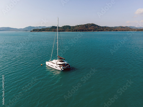 Aerial view of the white luxury yacht floating alone in the exotic turquoise sea near the island filled with forests and at the background of mountains and nature