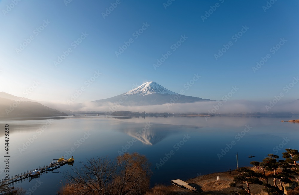 Morning scenery of snow capped Mount Fuji reflected in the peaceful lake water under blue clear sky with low clouds lingering at the foothill on a sunny winter day at Kawaguchiko, Yamanashi, Japan