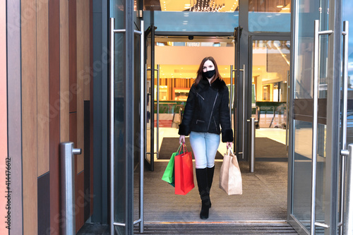 Beautiful woman in protective mask with shopping bags walks out mall. Stylish shopper girl