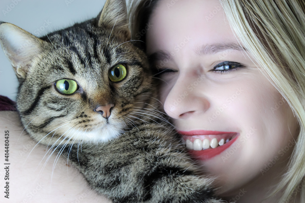 Young blonde woman smiles cheerfully hugging a gray tabby cat 