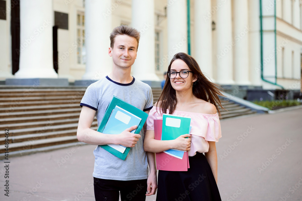 Two students standing together near the entrance of university, outdoors, looking to the camera. Two cheerful students, holding copybooks and folders.