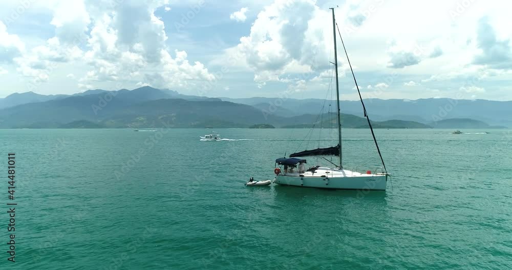 Aerial drone orbiting shot of moored sail boat in a turquoise calm ocean with people inside the boat getting it ready to sail in a beautiful tropical horizon with mountains Aerial drone orbiting shot of moored sail boat in a turquoise calm ocean with people inside the boat getting it ready to sail in a beautiful tropical horizon with mountains