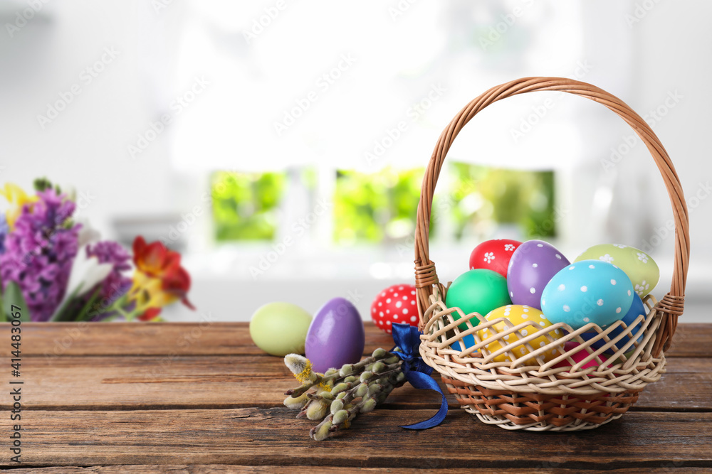 Colorful Easter eggs in wicker basket and willow branches on wooden table indoors, space for text