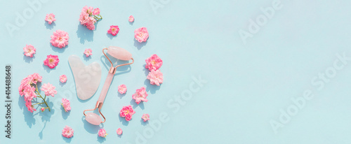 Facial massage kit for home spa. Face roller and gua sha massager made from rose quartz on blue pastel background with rose buds. Natural treatment concept.