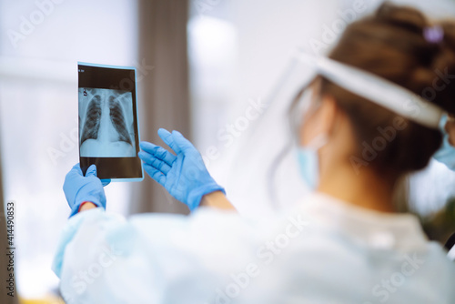 A female doctor in visor and protective gloves  discussing an x-ray. Doctor  examines an X-ray of a patient s lung infected with covid-19 coronavirus, pneumonia.X-rays of light. Fluorography. photo