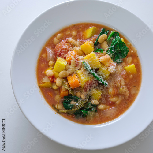 Ribollita or minestrone soup with beans, butternut squash, kale, bay leaf, and parmesan cheese served on the white shallow bowl. Diagonal view on white background, top view.