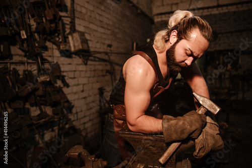 Muscular brutal blacksmith in a leather apron in the forge leaned on an anvil with a hammer © pantovich