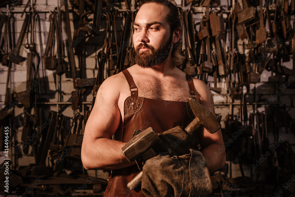 Muscular brutal blacksmith in a leather apron in the forge holding two hammers crossed in front of him Muscular brutal blacksmith in a leather apron in the forge holding two hammers crossed in front of him