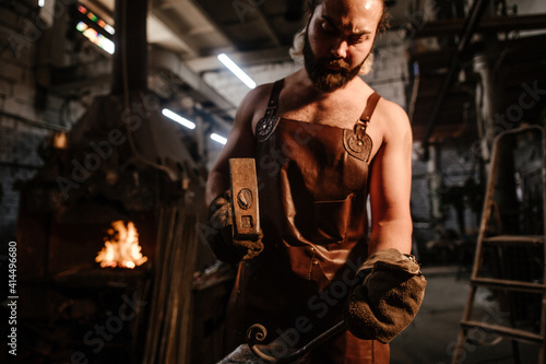 Blacksmith in a leather apron in a forge forging anvils with an iron bar © pantovich