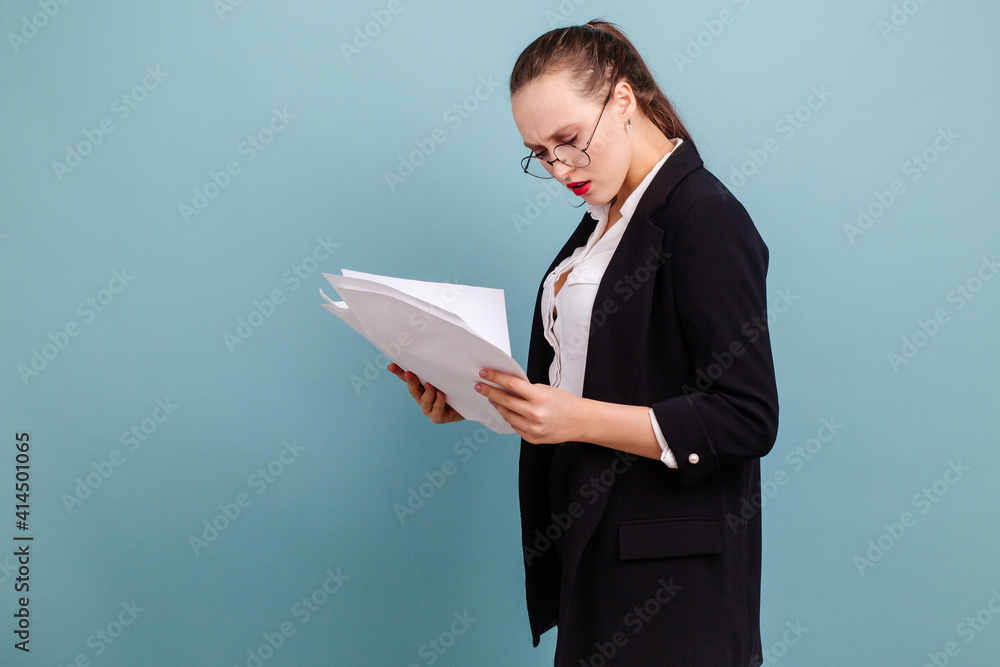 The girl with a dissatisfied look holding the pieces of paper looks at them standing on a blue background The girl with a dissatisfied look holding the pieces of paper looks at them standing on a blue background