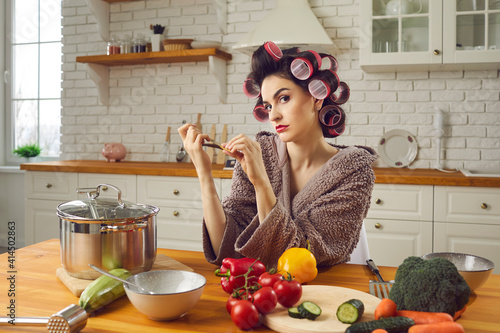 Woman in funny curlers sitting at kitchen table and filing nails with thoughtful face expression. Angry wife waiting for husband from work. Young housewife cooking, thinking and making cunning plans