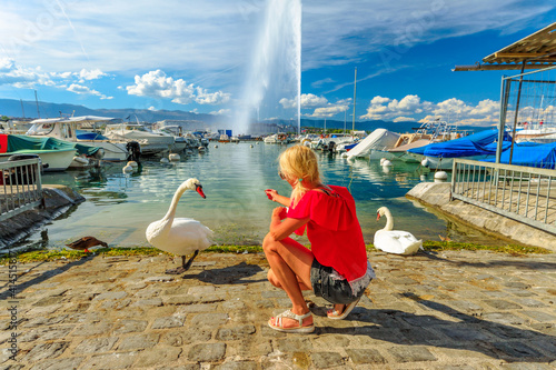 Blonde tourist girl feeding white swans on the shore of Geneva Lake in Geneva Harbor with 140m high fountain called Jet d'Eau. Marina with boats, sailboats and yachts. French Swiss, Switzerland.