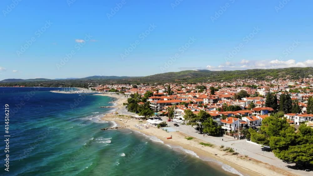 Aerial drone view of the Aegean sea coast in Nikiti, Greece. Beach with waves of the sea, umbrellas, sunbeds greenery along the embankment street, multiple buildings, hills in th distance Aerial drone view of the Aegean sea coast in Nikiti, Greece. Beach with waves of the sea, umbrellas, sunbeds greenery along the embankment street, multiple buildings, hills in th distance