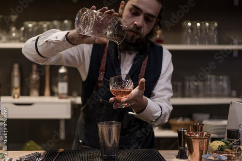 Barman in shirt and apron making an alcoholic drink with ice in a cocktail glass