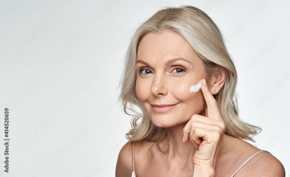 Smiling 50s middle aged mature older woman applying facial cream on face  looking at camera isolated on white background. Anti age healthy dry skin  care beauty therapy concept, old skincare treatment Stock