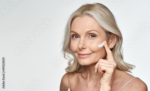 фотографія Smiling 50s middle aged mature older woman applying facial cream on face looking at camera isolated on white background