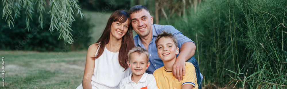 Happy Caucasian family of four people in park outdoor on summer day. Mother and father hugging two sons brothers siblings. Lifestyle authentic natural emotions moment. Web banner header. Happy Caucasian family of four people in park outdoor on summer day. Mother and father hugging two sons brothers siblings. Lifestyle authentic natural emotions moment. Web banner header.