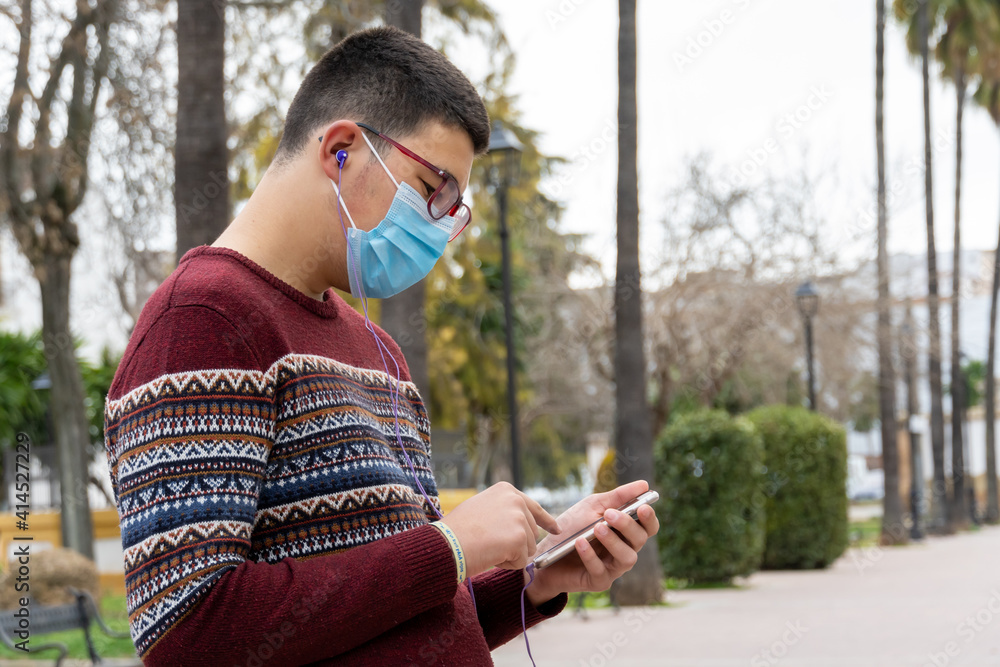 Stock photo of young boy wearing face mask due to covid19 using his phone and listening to music in the park. Stock photo of young boy wearing face mask due to covid19 using his phone and listening to music in the park.